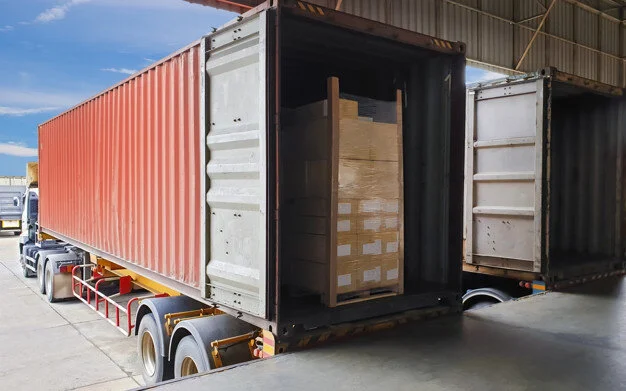 Red shipping container on a trailer at a loading dock, pallets stacked inside the open end.
