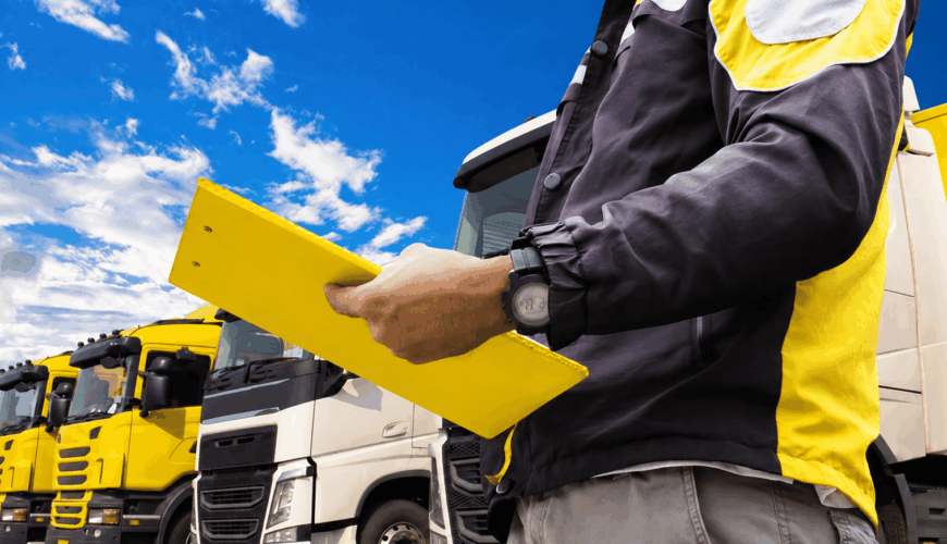 Worker in a high-visibility yellow vest inspecting with a yellow clipboard in front of a line of yellow trucks against a bright blue sky