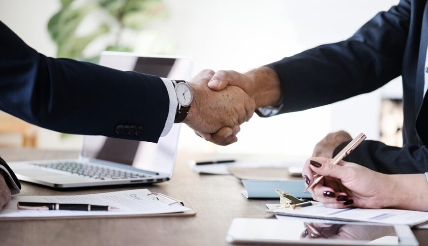 Two professionals shake hands across a desk, with a laptop, documents, and a smartphone visible in a business setting.