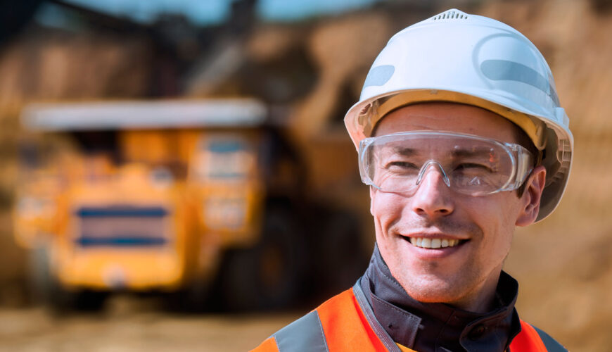 Construction worker wearing a white hard hat and clear safety goggles, smiling at a construction site with heavy equipment blurred in the background.