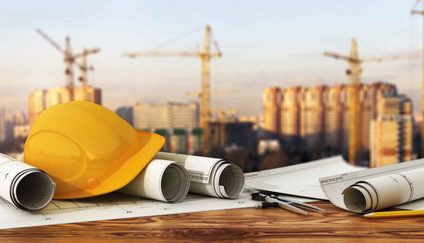 Yellow construction hard hat and rolled blueprints on a wooden desk with a blurred construction site in the background.