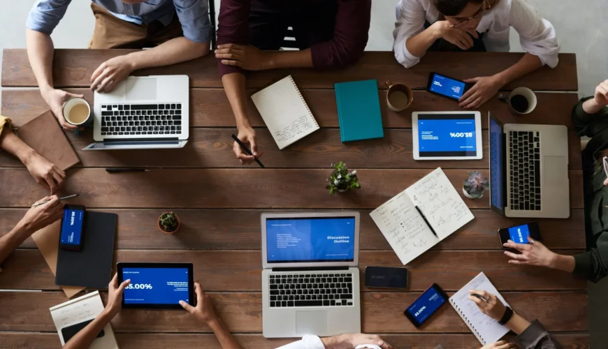 Overhead view of a diverse group gathered around a wooden table with laptops, tablets, phones, notebooks, and coffee cups, collaborating on a project.