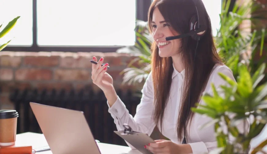 Smiling woman wearing a headset and holding a tablet while working at a desk with a laptop and plants nearby (customer support scene).