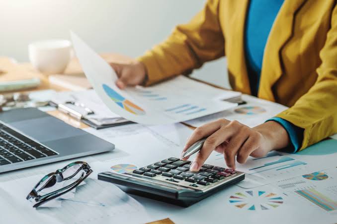 Person in a yellow blazer uses a calculator while reviewing financial charts and documents at a desk with a laptop nearby.